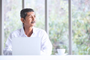 一位身穿白襯衫的中高齡男子坐在筆電前，望向窗外若有所思，桌上放著一杯咖啡，背景是模糊的綠色景致。