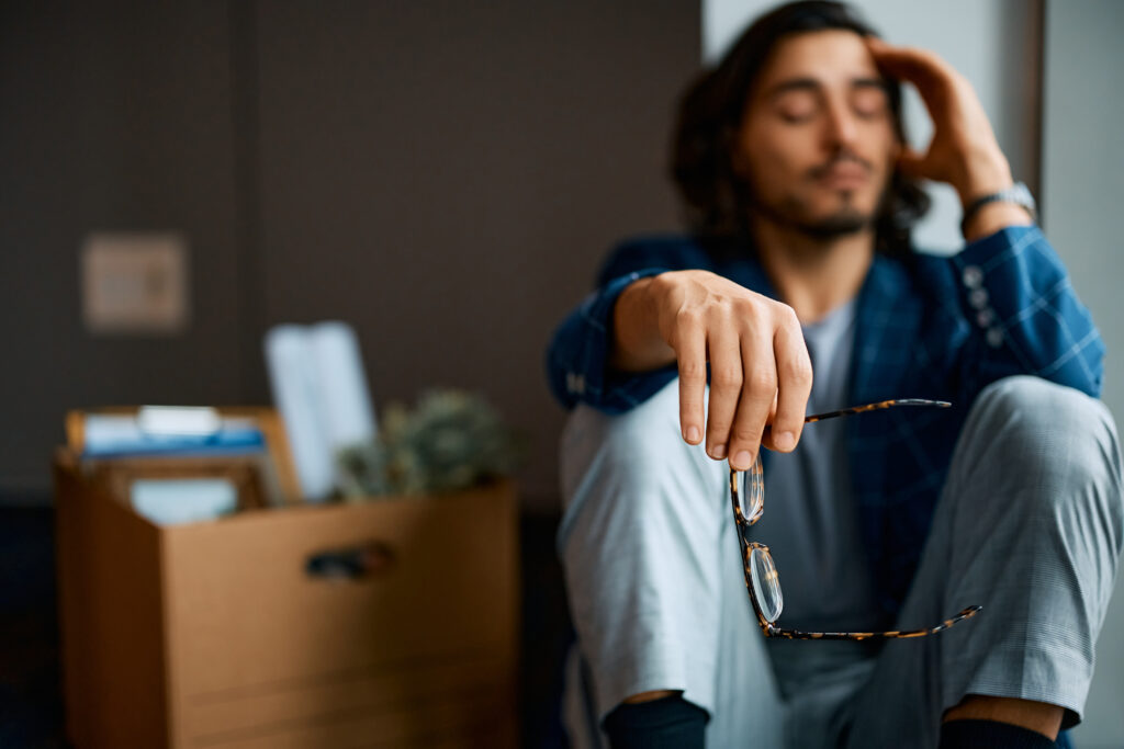 一位神情疲憊的男子坐在地上，手中拿著眼鏡，身旁有裝著文件和植物的紙箱，象徵失去工作後的無奈與迷惘。