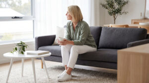 成熟女性坐在客廳沙發上,手持咖啡杯望向窗外,在自然光下安靜獨處