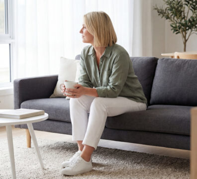 成熟女性坐在客廳沙發上,手持咖啡杯望向窗外,在自然光下安靜獨處